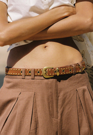 A close-up of a person wearing a brown studded leather belt with a gold-tone buckle, paired with a white top and pleated brown skirt.