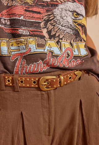 A close-up of a person wearing a brown graphic T-shirt with an eagle and car print, paired with a brown studded belt and brown pleated pants.
