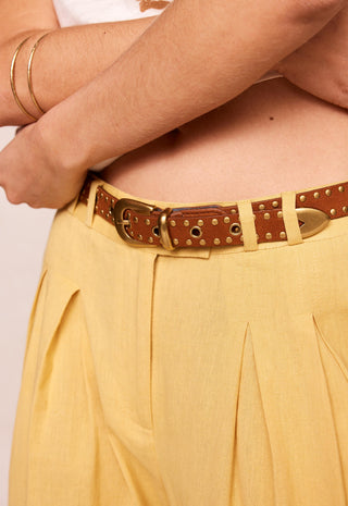 A close-up of a model wearing a brown leather belt with brass studs and a gold-tone buckle, paired with pleated yellow trousers.