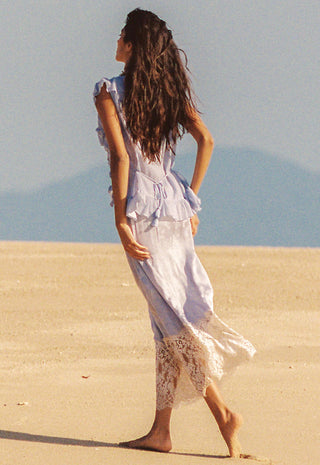 A woman with long dark hair stands barefoot on a sandy beach, viewed from behind, wearing a flowing light blue and white dress with lace trim.