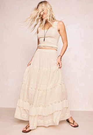 a-woman-modeling-a-cream-colored-two-piece-outfit-consisting-of-a-sleeveless-crop-top-and-a-long-tiered-maxi-skirt-with-delicate-lace-trim-paired-with-brown-flip-flop-sandals-against-a-textured-beige-studio-backdrop