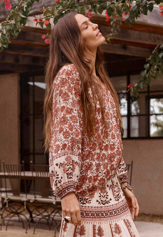 A woman wearing a bohemian-style long-sleeve dress with a rust-brown and cream floral pattern, standing in an outdoor patio setting with a rustic wooden structure and hanging greenery in the background.