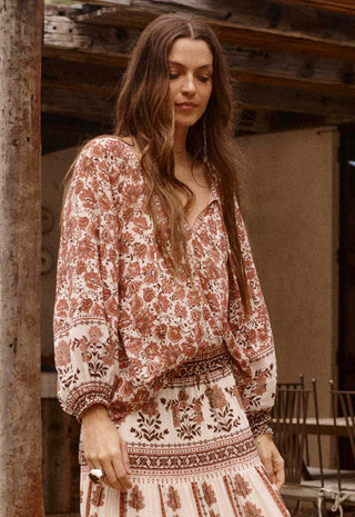 A woman wearing a bohemian-style long-sleeve maxi dress with a rust-brown and cream floral pattern, standing in front of a rustic wooden structure.