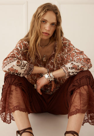 A woman wearing a bohemian-style brown and beige patterned blouse with sheer lace-trimmed sleeves and dark brown ruffled pants, seated against a white paneled wall.