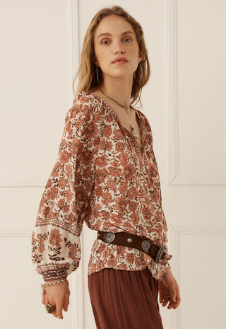 A woman wearing a bohemian-style long-sleeve blouse with a rust and beige floral print, paired with a dark brown belt and matching brown skirt, standing against a white paneled wall.