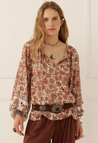 A woman modeling a long-sleeve bohemian-style blouse with a rust-brown and cream floral pattern, paired with a dark brown belt and brown pants, set against a white paneled wall.