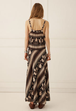 A woman viewed from behind wearing a sleeveless maxi dress with a brown, beige, and black striped and floral pattern, paired with brown flat shoes against a white paneled wall.