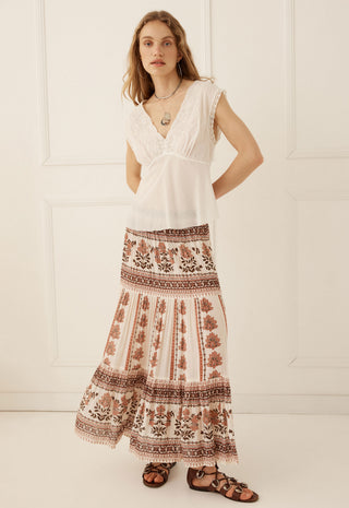A woman modeling a bohemian-style two-piece outfit featuring a white sleeveless lace-trimmed top and a long cream maxi skirt with brown floral and geometric patterns, paired with brown strappy sandals against a white paneled wall.