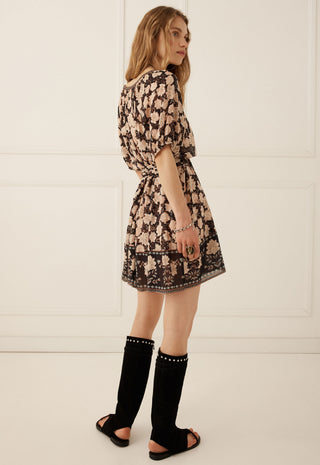A woman wearing a short black and beige floral print dress, styled with black knee-high studded sandals, standing against a white paneled wall.