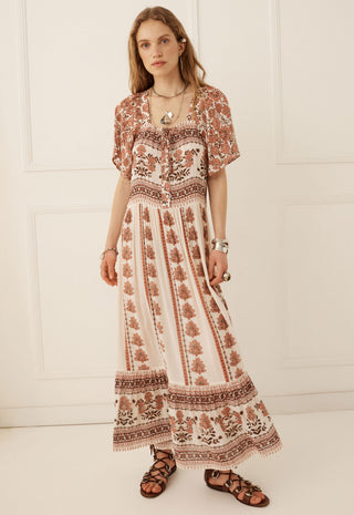 A woman modeling a bohemian-style maxi dress with a brown and beige floral pattern, standing against a white paneled wall.