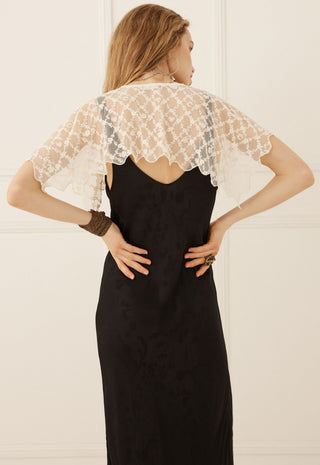A woman viewed from the back wearing a black slip dress with a sheer white lace flutter cape overlay, set against a light cream paneled wall.