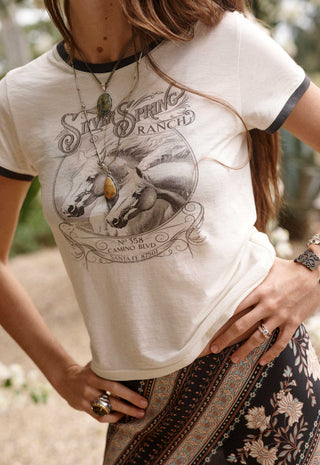A woman wearing a cream-colored ringer t-shirt with black trim and a vintage 'Silk Spring Ranch' graphic print on the front, paired with a dark patterned skirt.