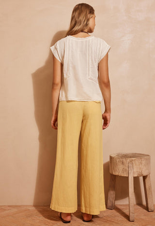 A woman stands with her back to the camera wearing a cream short-sleeve top and mustard yellow wide-leg pants, standing in front of a textured beige wall next to a rustic wooden stool.