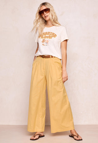 A woman modeling a white graphic t-shirt and yellow wide-leg pants with a brown belt, styled with sunglasses and flip-flops against a textured beige wall.