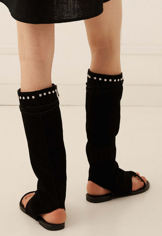 Black over-the-knee leg warmers with silver stud detailing at the top, worn by a model with black slide sandals on a light beige background.