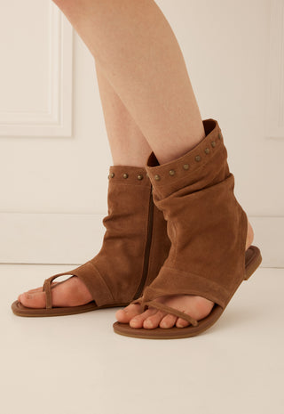 A pair of brown suede slide sandals with wraparound ankle straps and metal studs, shown on a model's feet against a light cream background.